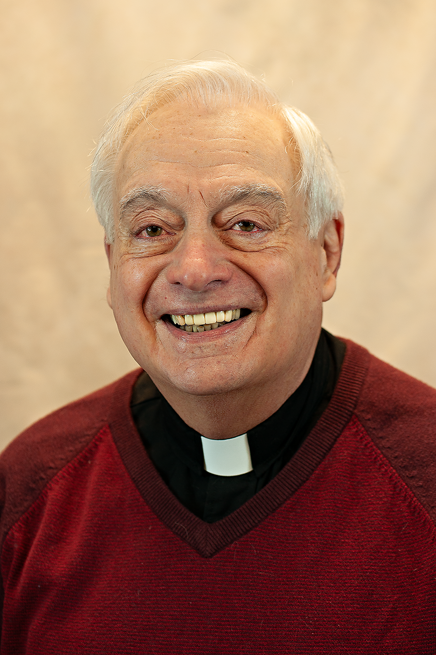 Rev. Nick Lorenzetti is an older priest wearing red with a priest's collar. He has white hair and is smiling.