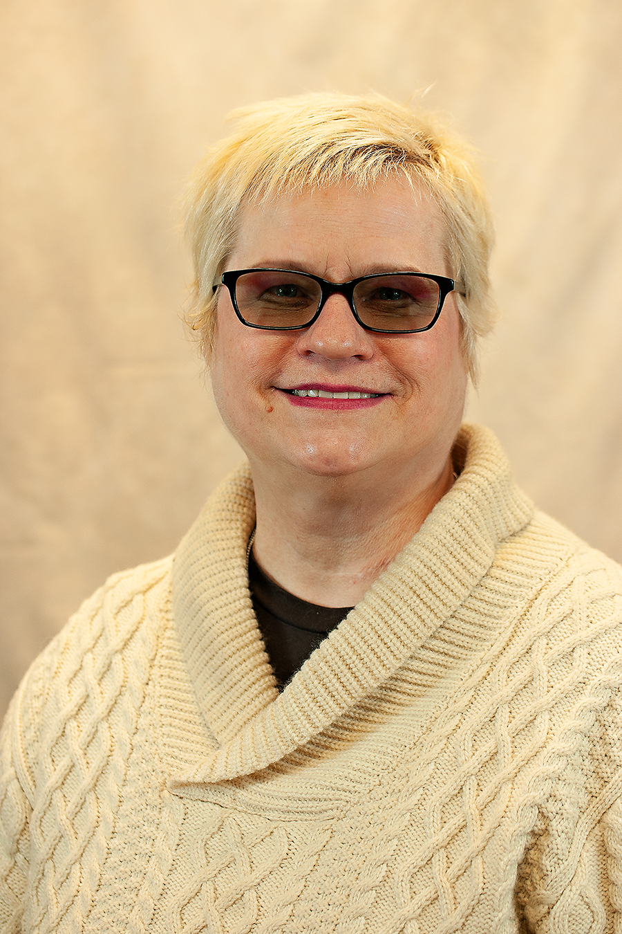 Middle-aged white woman with cropped blonde hair and dark glasses. She is wearing a fisherman's sweater. She is smiling.