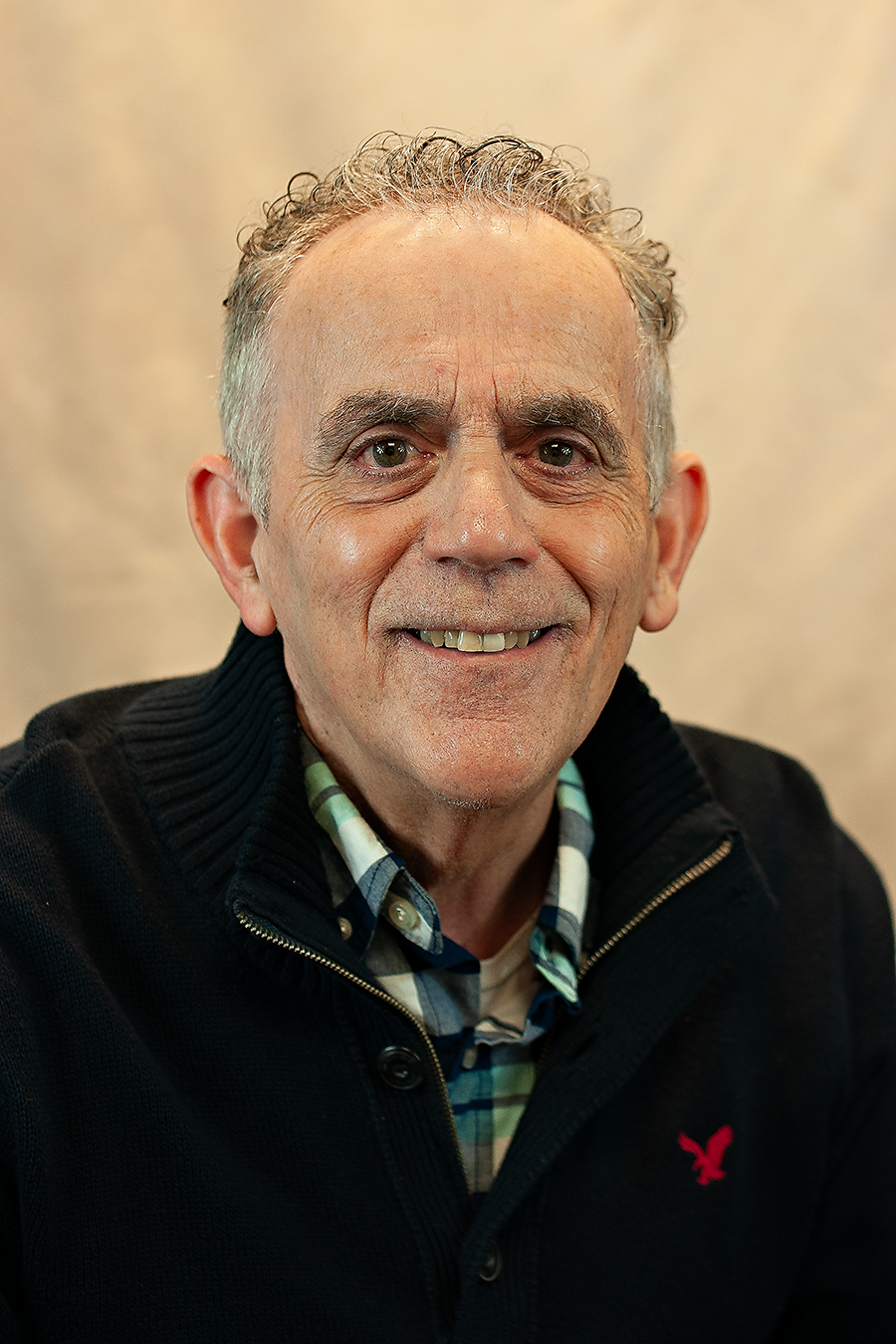 Older white man with grey hair, wearing a plaid shirt and a dark pullover. He is smiling.